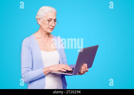 Gray haired old serious business woman wearing glasses, holding computer, typing texts, browsing. Isolated over violet purple background Stock Photo