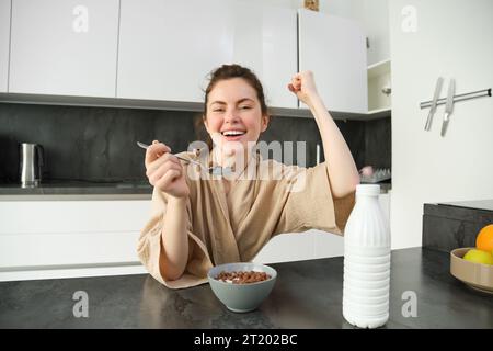 Portrait of enthusiastic young woman eating cereals with milk, looking excited and happy, sitting near kitchen worktop and having breakfast, raising h Stock Photo
