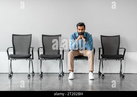 Long time of waiting. Thoughtful serious indian businessman holding hands on chin sitting on chair against grey wall Stock Photo