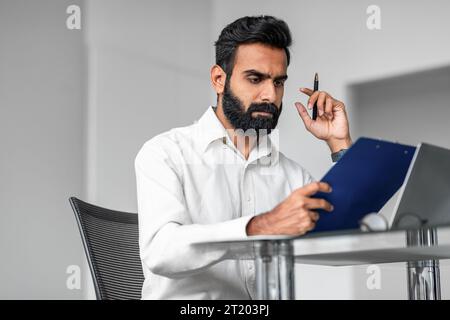 Serious indian bearded man sitting at table with laptop and clipboard, working with documents in office interior Stock Photo