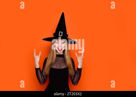 Photo portrait of lovely blonde teen lady showing horn sign hands rock dressed black halloween garment isolated on orange color background Stock Photo