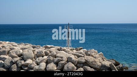 Metal beacon on rocky breakwater. Striped structure lighthouse, navigation sign on blue ripple sea background, sunny day. Stock Photo