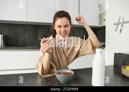 Portrait of enthusiastic young woman eating cereals with milk, looking excited and happy, sitting near kitchen worktop and having breakfast, raising h Stock Photo