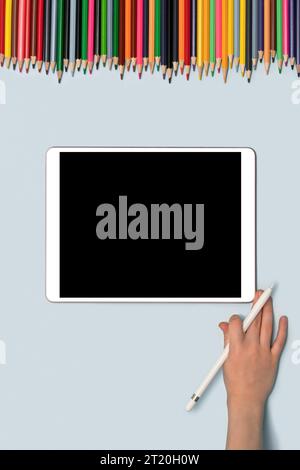 A tablet and a child's hand with a stylus are lying on the table, on top of colorful pencils. Concept of online education, lessons. Top view. Space fo Stock Photo