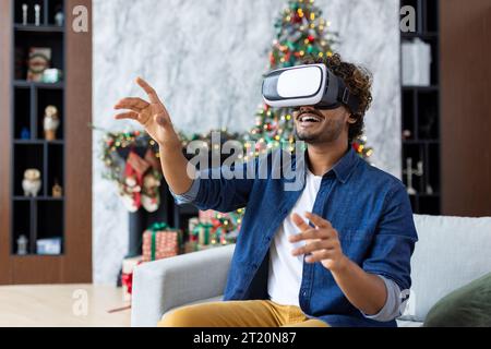 A man is using a virtual reality simulator for Christmas, sitting on a sofa in the living room of the house, near a decorated Christmas tree, on New Year's holidays. Stock Photo
