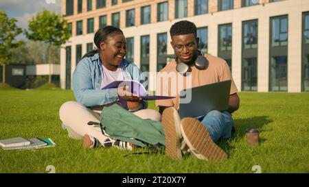 Two African American friends students high school in park studying together classmates university college outdoors learning with laptop on lawn girl Stock Photo