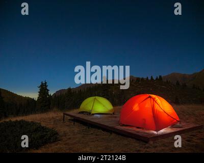 Tents are set up on a wooden platform in the mountains. Night time outdoors. Green and red tent with lighting inside Stock Photo