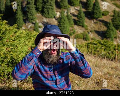 A bearded man looks through binoculars and is surprised. Outdoor Hiking Stock Photo