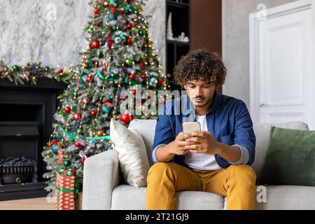 Serious thinking man is concentrating on choosing gifts and goods in an online store, hispanic sitting at home on sofa for Christmas, holding a smartphone, near the tree for New Year's holidays. Stock Photo