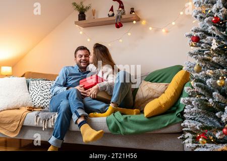 Christmas gift giving. A girl gives a gift to her boyfriend and kisses him on the cheek. Stock Photo
