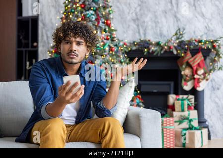 Portrait of dissatisfied upset man at home on Christmas and New Year holiday, hispanic looking disappointed at camera holding phone, sitting on sofa in living room. Stock Photo