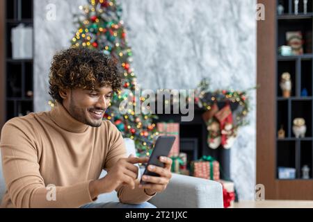 Young handsome hispanic man at home sitting on sofa in living room for Christmas near tree, man using phone happily smiling typing online text message and browsing internet pages. Stock Photo