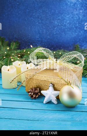 Golden yellow gift box with bow, burning candles, white star and pine cone on blue boards behind fir branch with blurred background. Christmas, New Ye Stock Photo