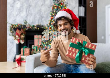 Online video call, holiday greeting for Christmas, man smiling and looking at camera in red Santa hat, sitting on sofa in living room, showing gift, Hispanic man greeting friends with New Year. Stock Photo