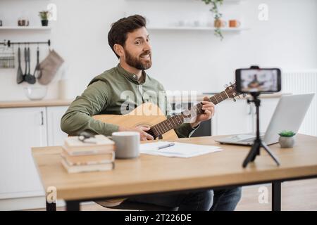 Skillful musician sitting at home office using smartphone and tripod for filming video tutorial about guitar playing. Handsome male blogger sharing wi Stock Photo