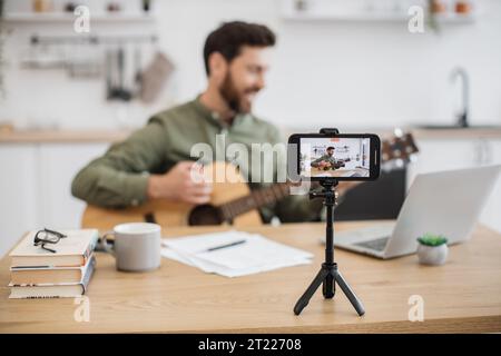 Skillful musician sitting at home office using smartphone and tripod for filming video tutorial about guitar playing. Handsome male blogger sharing wi Stock Photo