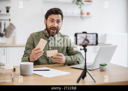 Emotional young guy opening cardboard box and looking inside with excitement while filming video on smartphone. Caucasian male blogger sharing positiv Stock Photo