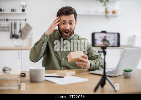 Emotional young guy opening cardboard box and looking inside with excitement while filming video on smartphone. Caucasian male blogger sharing positiv Stock Photo