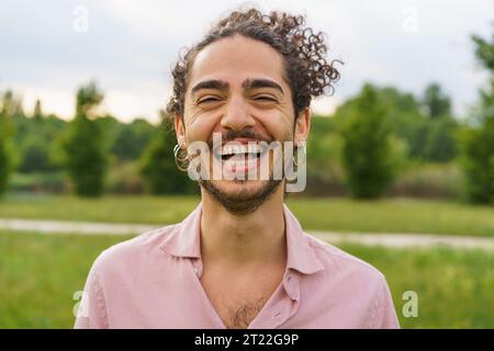 Outdoor portrait of a young man with curly hair tied back, laughing heartily with eyes squinted. He sports hoop earrings and a beard. The shot, taken Stock Photo
