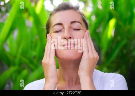 Close up photo of a middle-aged woman doing a facial massage using massage cream. Daily skin care and fight against age-related changes. Facial skin c Stock Photo