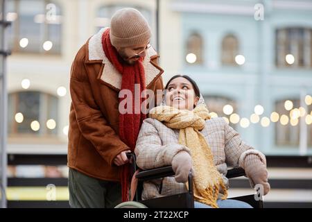 Portrait of young couple with smiling woman using wheelchair enjoying date outdoors in Christmas city Stock Photo