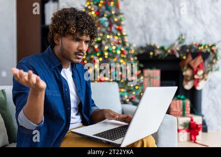 Upset man looking at laptop screen sitting at home in living room on Christmas, hispanic man frustrated writing online notification email to mail, near Christmas tree on New Year holidays. Stock Photo