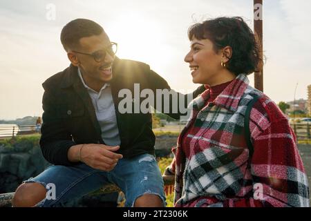 An African American young man sits on a railing, joking with his brunette friend. The two chat and smile at each other during a mid-season sunset, wit Stock Photo