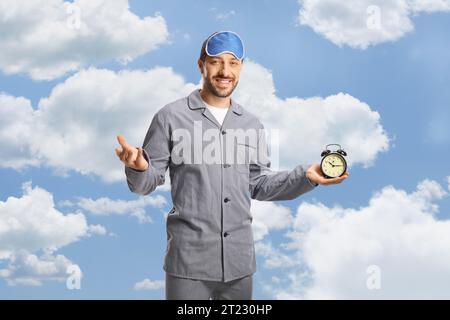 Young man in pajamas holding an alarm clock and smiling at camera with clouds and blue sky in the background Stock Photo