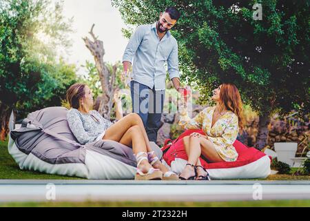 A bearded young man, around 25 years old, brings two cocktails to his friends who are engaged in a conversation while seated on bean bag chairs on the Stock Photo