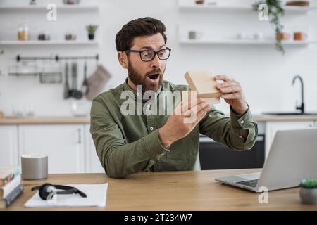 View from smartphone camera of emotional man in casual clothes experiencing pleasant shock while looking inside parcel box. Technology blogger satisfi Stock Photo