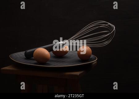Still life three eggs and an egg beater on a dark plate Stock Photo