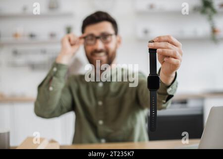 Blurred background of smiling bearded blogger sitting at home office and showing new purchased smart watch. Focus on new modern device in male hand. U Stock Photo