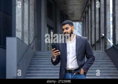 Young Indian man, real estate agent, broker standing near office outside and using phone, waiting for appointment, typing message. Stock Photo