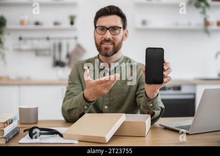 First person view of male influencer sharing feedback with followers while live streaming unpacking of modern smartphone. Bearded man sitting at desk Stock Photo