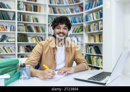 Portrait of a young Indian student studying online via video call, sitting in the library wearing headphones, writing an outline and smiling at the camera. Stock Photo