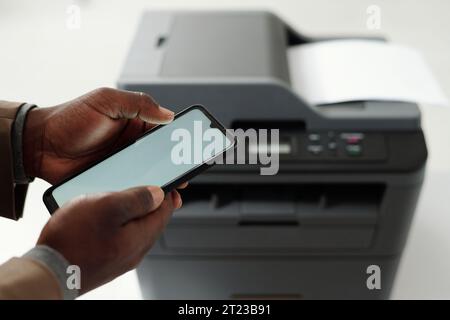 Screen of smartphone held by young African American businessman texting or scrolling through online data against xerox machine Stock Photo