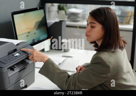 Young confident businesswoman pressing button on panel of xerox machine while setting number of copies of documents to make Stock Photo