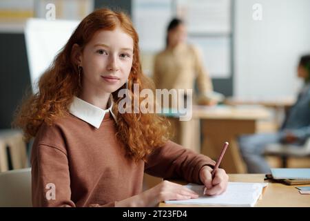 Youthful girl with pen in hand looking at camera while sitting by desk and writing down new rules of English grammar against teacher Stock Photo