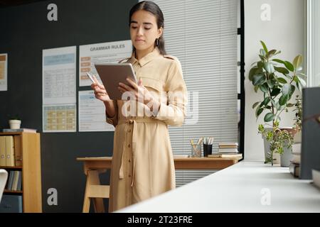 Young serious female teacher of English language in beige dress looking at tablet screen while watching educational online video in classroom Stock Photo