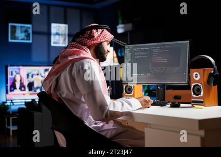 Dedicated software programmer wearing traditional Arabic clothing is seated at his home office desk examining database code on computer. Muslim man checking the programming language on the pc monitor. Stock Photo