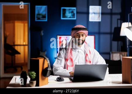 Confident Arab businessman working at a modern desk, typing on his laptop. Muslim guy engaged in browsing, email communication, and wireless connectivity in a calm and professional office setting. Stock Photo