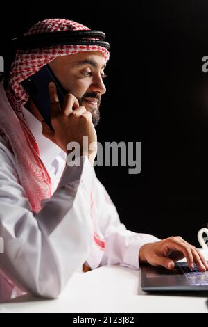 Middle Eastern man, dressed traditionally, engaged in a phone conversation, he multitasks with his mobile device and laptop. A modern blend of heritage and technology. Side-view, close-up. Stock Photo