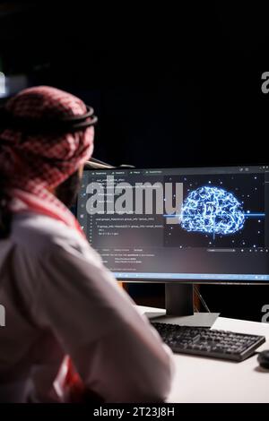 Muslim guy working with code and AI interface on a desktop computer. He is a professional developer managing data on a networked server in a cloud-based data center. Stock Photo