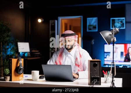 Focused man in Arabic attire working diligently at his modern office desk, typing on his laptop. Muslim guy surfing the net and checking his e-mail on his digital personal computer. Stock Photo