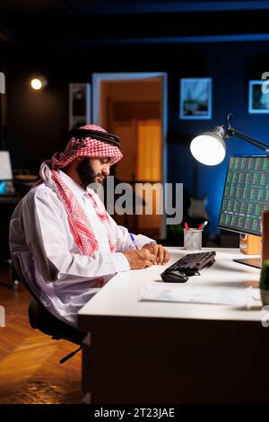Arab guy engrossed in his job, researching, taking notes, and surfing the net. By using his notepad and computer monitor, the professional Muslim man displays commitment and professionalism. Stock Photo