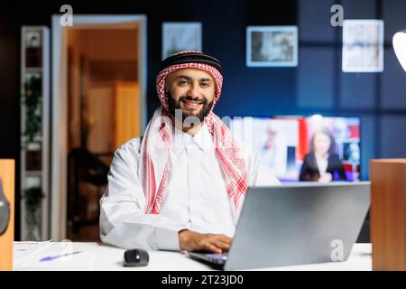Young man dressed in Arabian clothing using his laptop at the table. The Muslim guy, confident and tech-savvy, smiles at the camera, symbolizing the entrepreneurial mindset of the digital age. Stock Photo