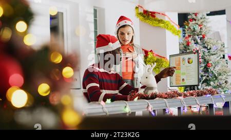 Employee working in Christmas ornate office asking colleague for help after getting stuck on task. Woman with Santa hat listening to coworker explaining problem while looking over financial figures Stock Photo