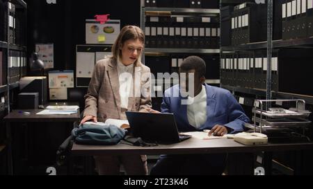 African american and caucasian police officers conduct thorough investigations and examine evidence files. Multiracial private detectives accurately cross-checking witness statements and clues. Stock Photo