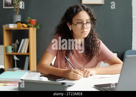 Serious learner of secondary school looking at laptop screen and making notes in copybook while listening to teacher explaining new topic Stock Photo