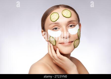 Closeup portrait of smiling attractive beautiful woman making skin care procedures, applying under eyes patches and cucumbers on face. Indoor studio shot isolated over gray background. Stock Photo